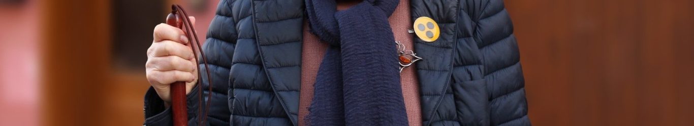 Man sieht eine Person in einer blauen Steppjacke mit einem Blinden-Symbol-Buttton. In der Hand hält sie einen Langstock.